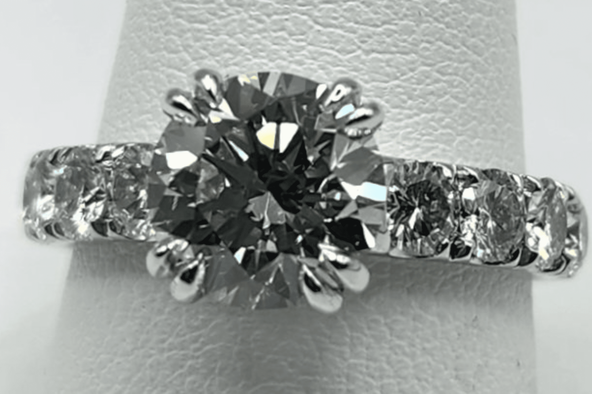 A diamond ring with a large central stone and smaller side diamonds on a white stand (1) A diamond ring with a large central stone and smaller side diamonds on a white stand
